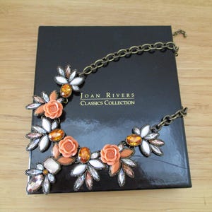 May include: A gold chain necklace with a statement piece featuring orange and white flowers with rhinestones. The necklace is displayed on a black box with the text "JOAN RIVERS CLASSICS COLLECTION".