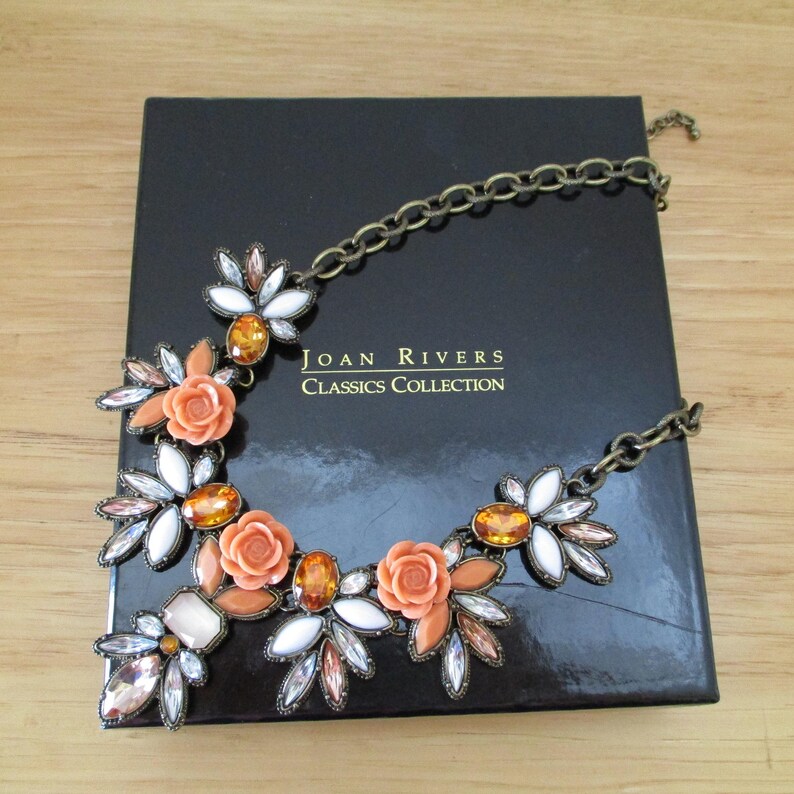 May include: A gold chain necklace with a statement piece featuring orange and white flowers with rhinestones. The necklace is displayed on a black box with the text "JOAN RIVERS CLASSICS COLLECTION".