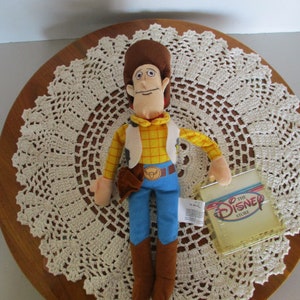 Puede incluir: Un peluche del personaje Woody de la pel&iacute;cula de Disney Toy Story. Woody lleva una camisa a cuadros amarilla, un chaleco, pantalones azules y botas marrones. El juguete tiene una etiqueta de The Disney Store.