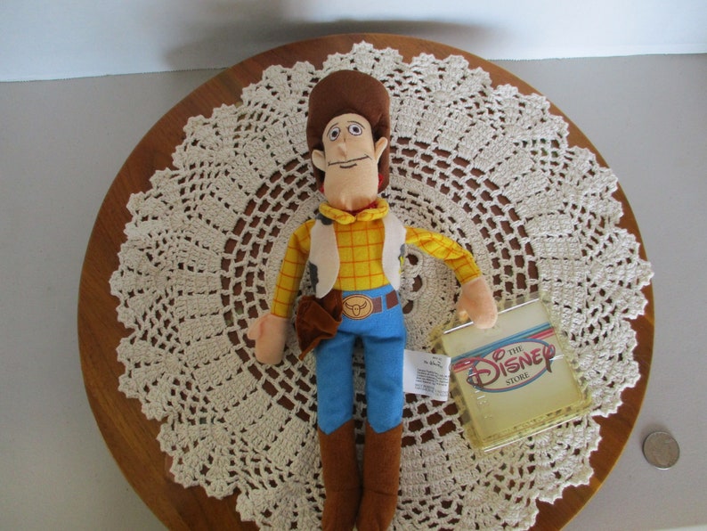 Puede incluir: Un peluche del personaje Woody de la pel&iacute;cula de Disney Toy Story. Woody lleva una camisa a cuadros amarilla, un chaleco, pantalones azules y botas marrones. El juguete tiene una etiqueta de The Disney Store.