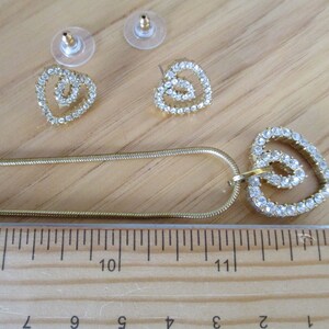 Puede incluir: Un collar de cadena dorada con un colgante de coraz&oacute;n doble y pendientes a juego. Los corazones est&aacute;n hechos de diamantes de imitaci&oacute;n transparentes.