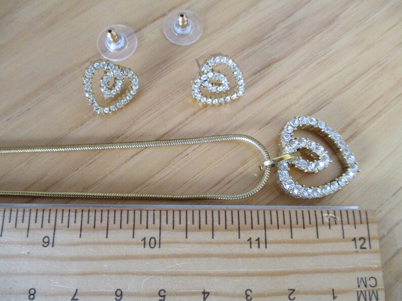 Puede incluir: Un collar de cadena dorada con un colgante de coraz&oacute;n doble y pendientes a juego. Los corazones est&aacute;n hechos de diamantes de imitaci&oacute;n transparentes.