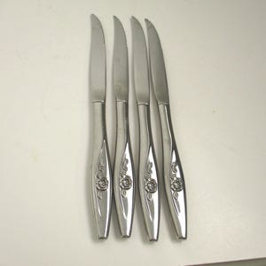 May include: A set of four silver-toned stainless steel steak knives with serrated blades. Each handle features a raised, embossed floral rose design with leaves, extending from the base of the blade down to the bottom of the handle.