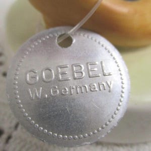 May include: Close-up of a silver metal tag with the embossed text "GOEBEL W. Germany". The tag is round with a beaded edge and a hole at the top for attachment. The background is blurred, suggesting a figurine or other object.