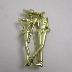 May include: Gold-toned metal brooch featuring a stylized couple, a woman in a hat and a man in a suit, standing side-by-side.