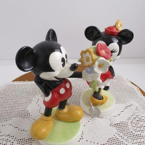 May include: A pair of ceramic figurines depicting Mickey Mouse and Minnie Mouse. Mickey, in red shorts and yellow shoes, extends his arm towards Minnie, who holds a bouquet of flowers. Both figures stand on a green base.