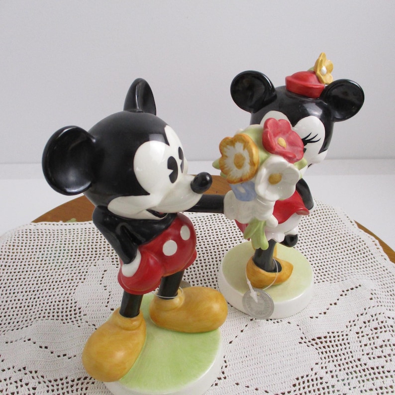 May include: A pair of ceramic figurines depicting Mickey Mouse and Minnie Mouse. Mickey, in red shorts and yellow shoes, extends his arm towards Minnie, who holds a bouquet of flowers. Both figures stand on a green base.