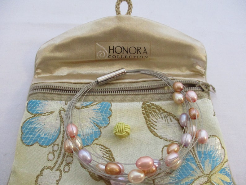 Op de afbeelding: Een meerstrengs parelketting met een magneetsluiting, in een beige satijnen zakje met de tekst "Honora Collection". Het zakje heeft een bloemenpatroon met blauwe en groene accenten.