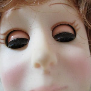 Puede incluir: Primer plano de la cara de una mu&ntilde;eca con los ojos cerrados y las mejillas sonrosadas. La mu&ntilde;eca tiene el pelo casta&ntilde;o y rasgos realistas.