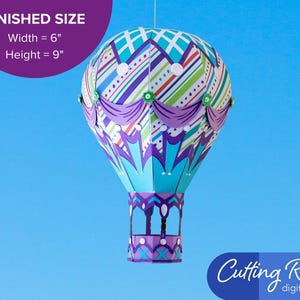 A papercraft hot air balloon with a striped pattern in shades of blue, green, purple, and white. The balloon has purple swag accents and silver embellishments.  A banner indicates that the balloon is about 6 inches wide and 9 inches tall.