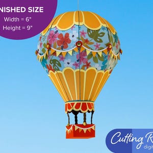 A papercraft hot air balloon with a blue and yellow color scheme. The balloon is decorated with a floral pattern and yellow and white bunting. A banner indicates that the balloon is about 6 inches wide and 9 inches tall.