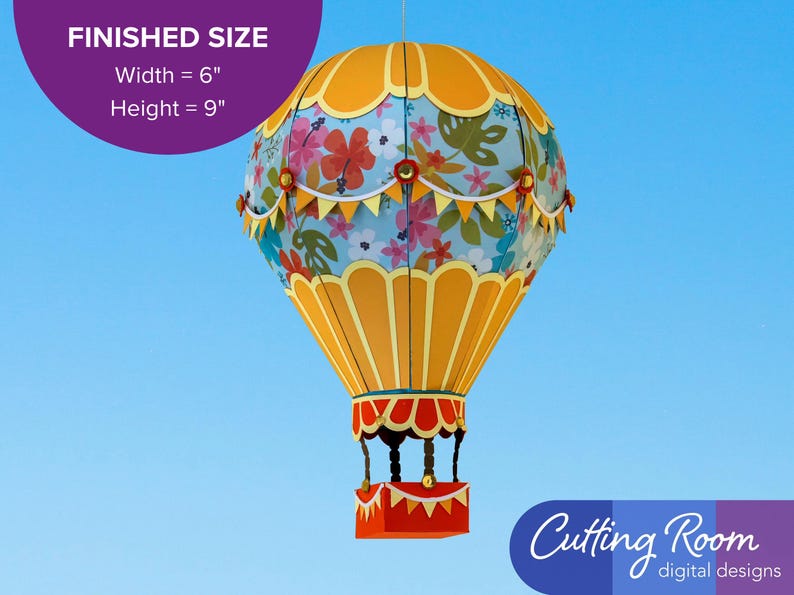 A papercraft hot air balloon with a blue and yellow color scheme. The balloon is decorated with a floral pattern and yellow and white bunting. A banner indicates that the balloon is about 6 inches wide and 9 inches tall.