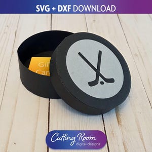 May include: Black round gift box with a hockey puck design on the lid. The lid features a gray circle with crossed hockey sticks. The box is open, revealing a yellow gift card inside. The text "SVG + DXF DOWNLOAD" is at the top.