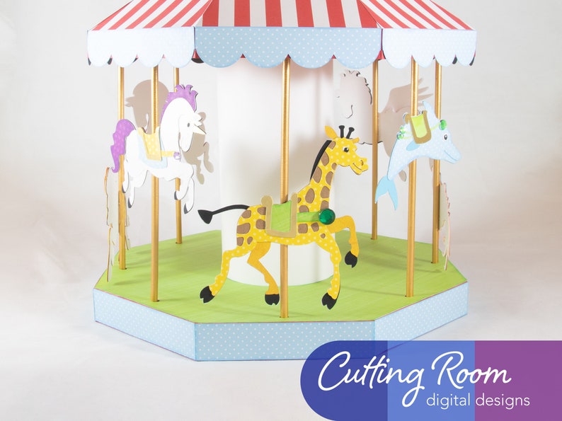 A papercraft carousel with a red and white striped roof, a blue and white polka dot base, and a white center pole. The carousel has four animals: a white horse with a purple mane, a yellow giraffe, a blue dolphin, and a white horse with a brown mane. The carousel is on a white background. Cutting Room digital designs.