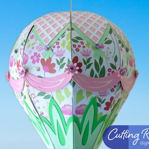A papercraft hot air balloon with a pink, green, and white floral pattern. The balloon has pink and green trim and a pink and white checkered top. The balloon is hanging from a string. The text 'Cutting Room digital designs' is in the lower right corner of the image.