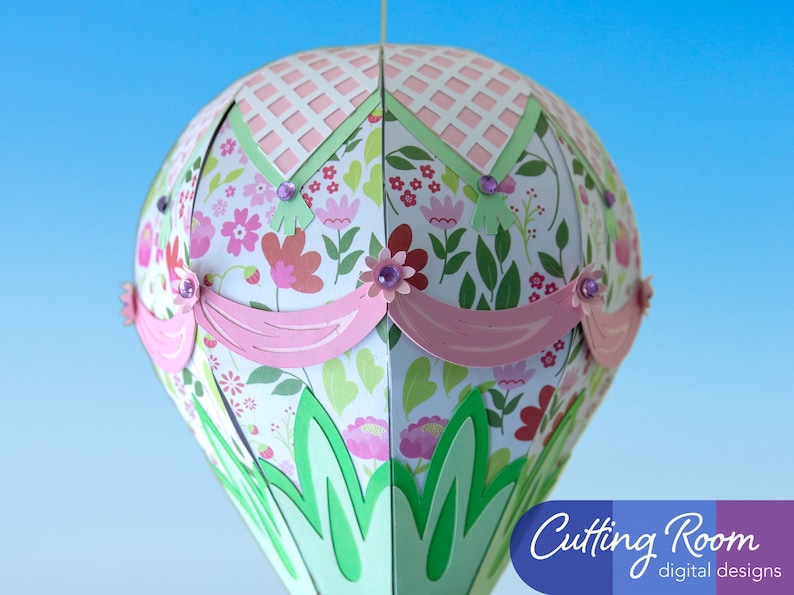 A papercraft hot air balloon with a pink, green, and white floral pattern. The balloon has pink and green trim and a pink and white checkered top. The balloon is hanging from a string. The text 'Cutting Room digital designs' is in the lower right corner of the image.