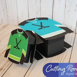 Two hockey jersey shaped boxes, one green and one blue, with black hockey sticks displayed on the front of each. The boxes are made of paper and are designed to be assembled. The text 'Cutting Room digital designs' is printed on a blue, purple, and white background at the bottom of the image.