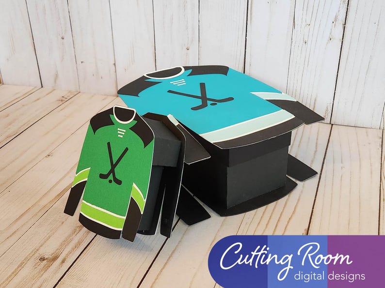 Two hockey jersey shaped boxes, one green and one blue, with black hockey sticks displayed on the front of each. The boxes are made of paper and are designed to be assembled. The text 'Cutting Room digital designs' is printed on a blue, purple, and white background at the bottom of the image.