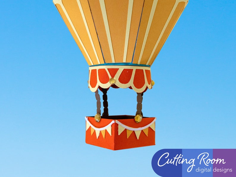 A papercraft hot air balloon with an orange basket. The balloon is made of orange and yellow paper with white stripes. The balloon is against a blue sky background.