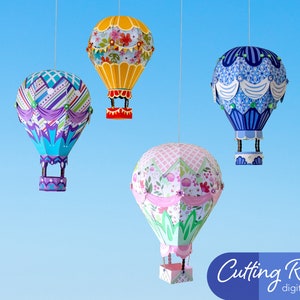 Four paper hot air balloons hanging from strings against a blue background. The balloons are decorated with different patterns and colors. The balloons are all different sizes. The balloons are labeled 'Cutting Room digital designs'.