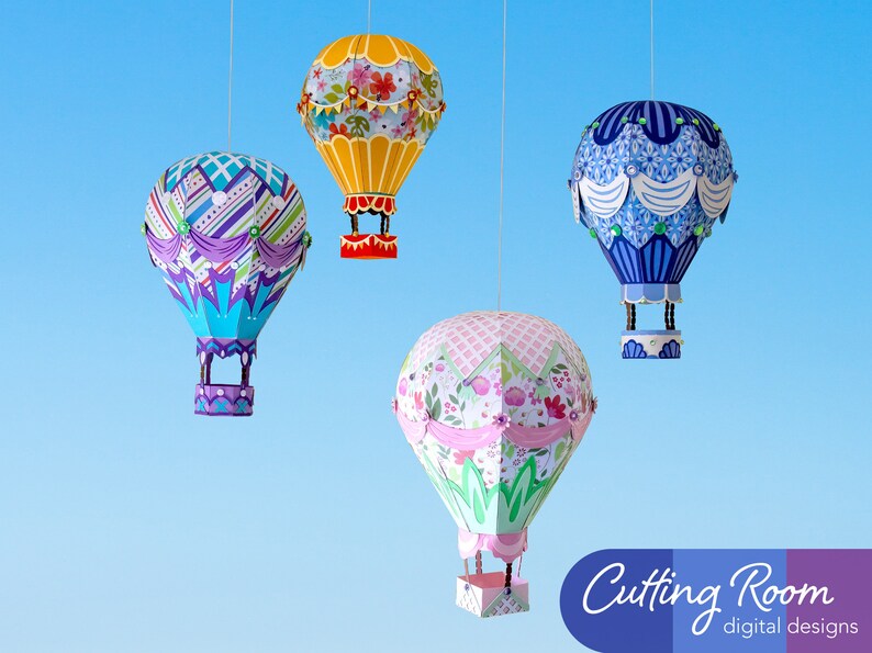 Four paper hot air balloons hanging from strings against a blue background. The balloons are decorated with different patterns and colors. The balloons are all different sizes. The balloons are labeled 'Cutting Room digital designs'.