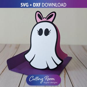May include: A white ghost-shaped paper craft with a pink bow and eyelashes, set on a purple envelope. The design includes the text "SVG + DXF DOWNLOAD" and "Cutting Room digital designs".