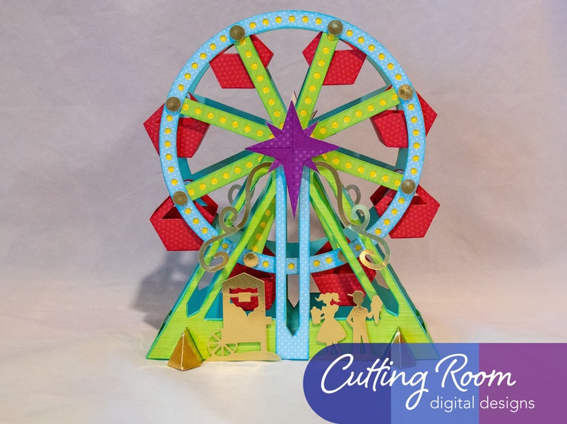 A papercraft model of a Ferris wheel with red and blue gondolas, green supports, and a purple star at the top. The model is on a green base with a gold cart and two figures standing in front of it.