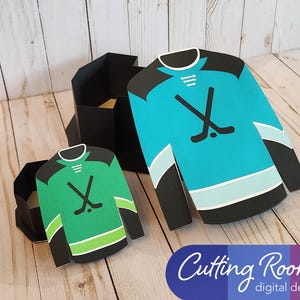 May include: Two hockey jersey shaped boxes, one green and one blue, with black trim and hockey stick and puck designs. They are sitting on a wooden surface and are open so you can see a little inside each box.