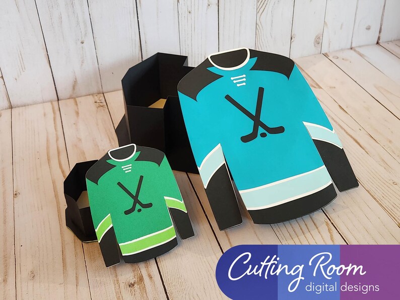 May include: Two hockey jersey shaped boxes, one green and one blue, with black trim and hockey stick and puck designs. They are sitting on a wooden surface and are open so you can see a little inside each box.