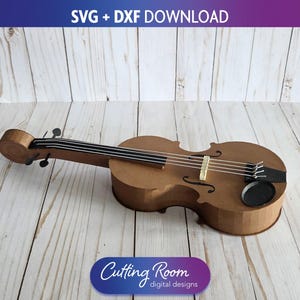 Puede incluir: Un violín de papel marrón con cuerdas y clavijas negras. El violín es un modelo 3D, probablemente para manualidades o diseño. La imagen incluye el texto "SVG + DXF DOWNLOAD" y "Cutting Room digital designs."