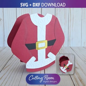 Puede incluir: Un traje de Papá Noel de papel rojo con ribetes blancos y una hebilla dorada, junto con una caja más pequeña a juego. El diseño es para una descarga digital, con el texto "SVG + DXF DOWNLOAD" en la parte superior y "Cutting Room digital designs" en la parte inferior.