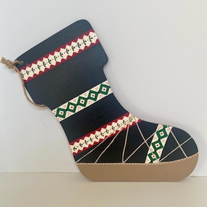 May include: A wooden Christmas stocking decoration. The stocking is black with red, green, and white patterned bands. The toe is a light brown color. A jute string is attached for hanging.