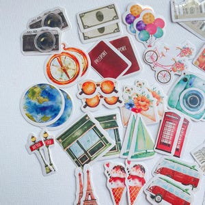 Op de afbeelding: Een verzameling reisstickers met illustraties van camera's, bankbiljetten, ballonnen, een kompas, paspoorten, zonnebrillen, een fiets, een wereldbol, bloemen, een telefooncel, een zeilboot, een metro-bord, een winkelpui, ijsjes en een busje.