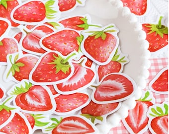 Puede incluir: Una colección de pegatinas de fresa rojas y verdes. Las pegatinas están dispuestas en un plato blanco con un borde de cuentas. El plato está sobre un fondo a cuadros rosa y blanco.