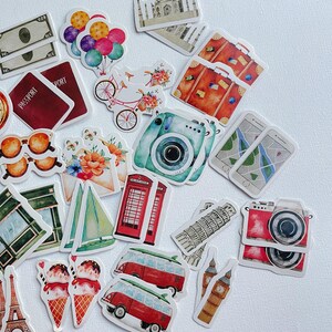 Op de afbeelding: Een verzameling stickers met reisthema met illustraties van een rode telefooncel, een blauwgroene camera, een rode camera, koffers, paspoorten, bankbiljetten, een fiets, een zeilboot, ijshoorntjes, een kaart op een telefoon en bezienswaardigheden.