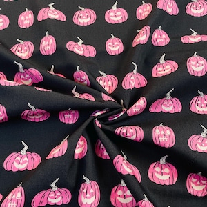 Op de afbeelding: Zwarte stof met een herhaald patroon van roze pompoenen. Elke pompoen heeft een lachend gezicht en een groene steel. De pompoenen variëren in grootte en oriëntatie, wat een speels Halloween-thema creëert.