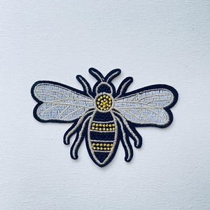 Op de afbeelding: Een geborduurde bij-patch in zwart en geel met witte vleugels. De bij heeft een gedetailleerd ontwerp met gele strepen en een zwart lichaam.