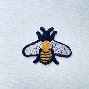 Op de afbeelding: Een geborduurde bij-patch in zwart en geel met witte vleugels en rode stiksels.