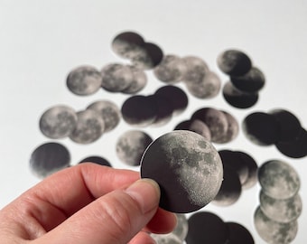 Op de afbeelding: Een verzameling ronde stickers met een gedetailleerde fotografische afdruk van de maan in verschillende fasen. Elke sticker toont een contrastrijke zwart-witafbeelding van het maanoppervlak met kraters en texturen tegen een donkere achtergrond.