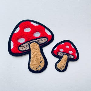 Op de afbeelding: Twee rode en witte, paddenstoelvormige viltpleisters met bruine stelen. De grotere paddenstoel heeft witte stippen op de rode dop.