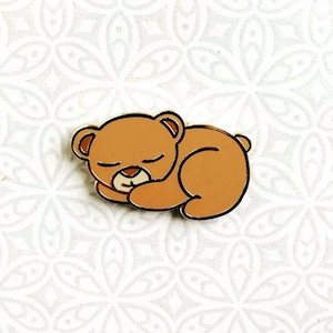 May include: A brown bear pin with closed eyes, curled up in a sleeping position. The bear is outlined in gold and has a gold nose.