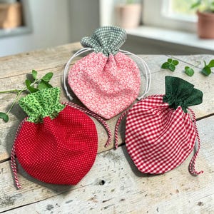 May include: Three fabric drawstring pouches shaped like strawberries. One is red with white polka dots and a green leaf top, one is pink with a white floral pattern and a green gingham top, and one is red and white gingham with a dark green leaf top.