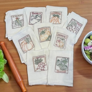 May include: A collection of white embroidered kitchen towels featuring various vegetables. Each towel displays a different vegetable, including corn, eggplant, carrots, beets, pumpkin, squash, peppers, radishes, and broccoli, with the vegetable's name embroidered below.