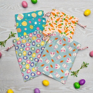 May include: Four drawstring fabric bags with Easter-themed patterns. One bag is turquoise with egg designs, another has carrot patterns, a third features eggs and flowers, and the last has bunny and carrot prints. Colorful Easter eggs are scattered around.