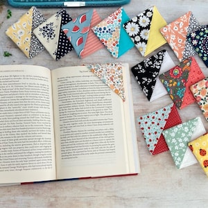 May include: A collection of ten handmade fabric bookmarks in various patterns and colors. The bookmarks are folded into triangles and feature a variety of designs, including floral, geometric, and polka dots.