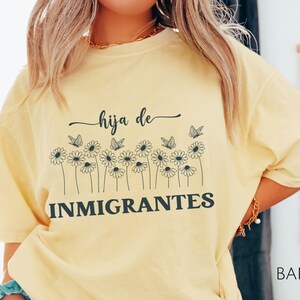 May include: A yellow t-shirt with a graphic design featuring a row of stylized daisies with butterflies and the text "hija de inmigrantes".