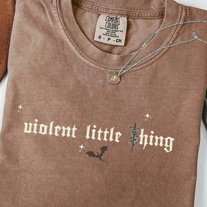 May include: A brown Comfort Colors t-shirt with the phrase "violent little thing" printed on the front. The shirt has a crew neck and short sleeves. A silver necklace with a small pendant is on the shirt.