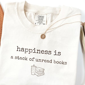 May include: Ivory-colored t-shirt with the text "happiness is a stack of unread books" in black. A small graphic of a coffee cup and books is below the text. A gold necklace with a pendant is draped on the shirt.