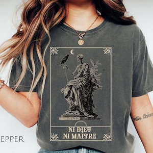 May include: Pepper-colored t-shirt featuring a detailed graphic of a skeleton holding a raven, with a crescent moon and floral elements. The text "NI DIEU NI MAITRE" is printed below the graphic. The shirt is a comfortable, casual style.