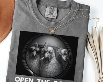 May include: Pepper-colored t-shirt with a black and white graphic of three chickens, one wearing sunglasses and another with a cigarette. The text "OPEN THE DOOR" is printed below the graphic. A silver necklace with a pendant is on the shirt.